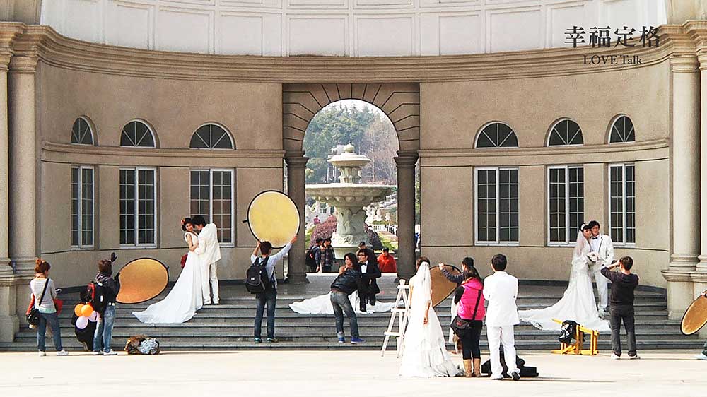 四對新人分散站在羅馬式建築物前拍攝婚紗。有白紗與黑西裝，也有白紗與白西裝，攝影師們和助理們專心地為新人們拍攝，記下這珍貴的、一生也許只有一次的時刻。大家都笑容滿面，是幸福洋溢的氛圍。